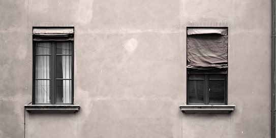 Two windows are set in a plain, gray concrete wall. The window on the left has partially open blinds with visible white curtains, while the window on the right is covered with a wrinkled, closed blind. The facade appears slightly weathered, creating a minimalistic and somewhat industrial look.