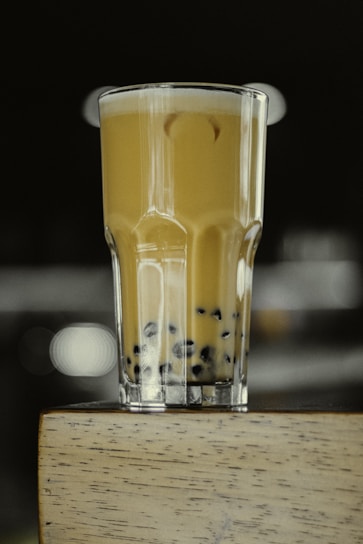 A tall glass of milk tea with black tapioca pearls settled at the bottom, placed on a wooden surface. The drink is creamy with a smooth texture, and the background is blurred with soft lighting.