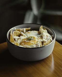 A bowl filled with a dish featuring tortilla chips covered in creamy sauce and topped with a perfectly cooked fried egg. Fresh herbs are sprinkled on top, adding a touch of green. The setting is cozy with a wooden surface beneath the bowl, and the background is softly blurred.