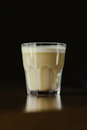A glass of layered coffee with a frothy top sits on a reflective surface against a dark background. The lighting focuses on the coffee, creating a warm and inviting ambiance.
