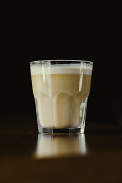 A glass of layered coffee with a frothy top sits on a reflective surface against a dark background. The lighting focuses on the coffee, creating a warm and inviting ambiance.