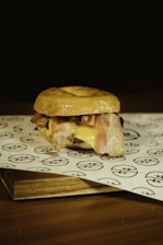 A sandwich is placed on a piece of branded parchment paper. It consists of a bun, slices of crispy bacon, melted cheese, and possibly other fillings. The background is dark, highlighting the sandwich as the focal point.