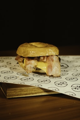A sandwich is placed on a piece of branded parchment paper. It consists of a bun, slices of crispy bacon, melted cheese, and possibly other fillings. The background is dark, highlighting the sandwich as the focal point.