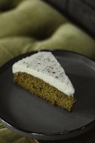 A slice of cake with a light brown base and a thick layer of cream or frosting on top, served on a dark plate. The frosting is sprinkled with a small amount of spice or cocoa powder. The background features a soft, blurred cushion, adding an element of comfort to the scene.