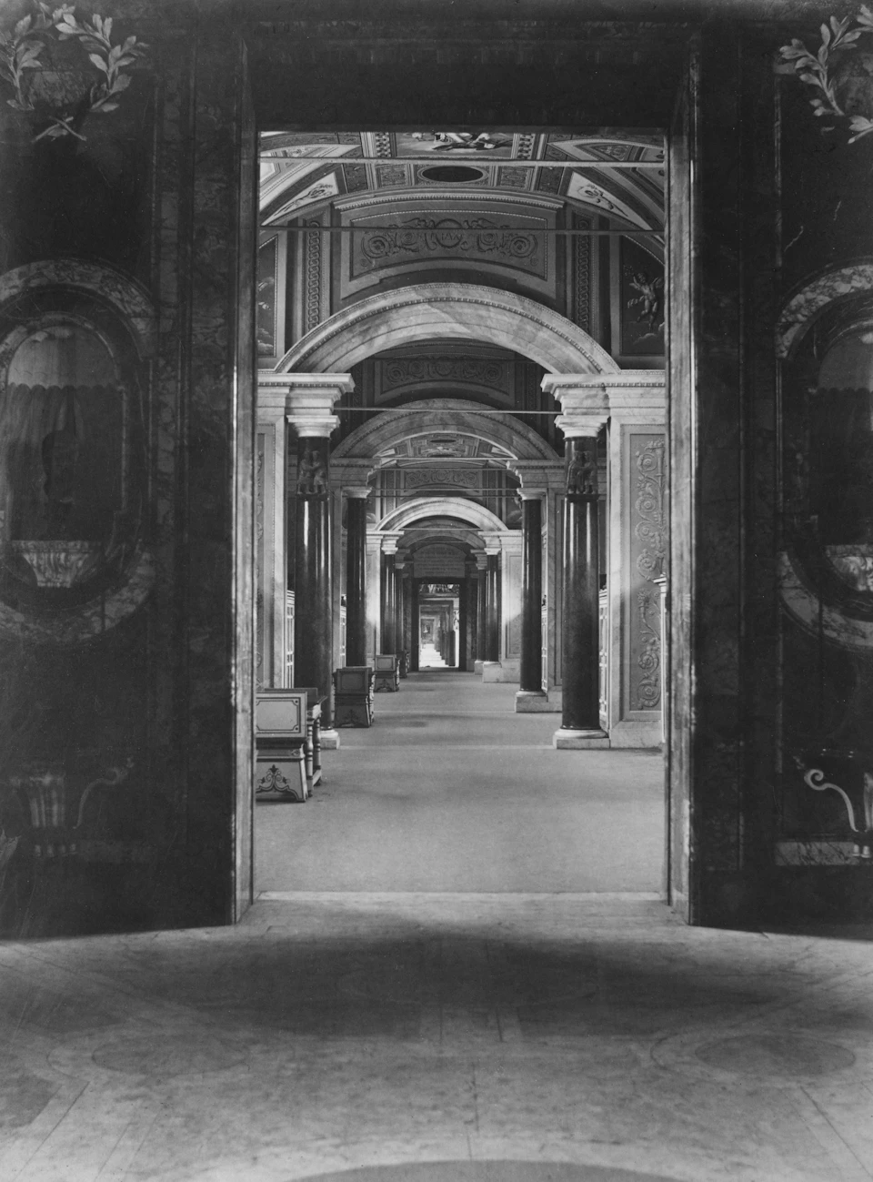 The image depicts a grand, long hallway with numerous columns lining both sides. The ceiling is adorned with intricate designs and patterns, adding to the overall elegance of the space. The perspective of the photo is from within one of the columns, looking out onto the hallway. The hallway itself appears to be empty at the moment, creating an atmosphere of tranquility and timelessness.