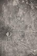 A detailed grayscale image of a lunar surface featuring numerous craters and rugged terrain. The surface is pocked with a variety of circular depressions and raised areas, characteristic of an impact-rich environment. Textured patterns radiate from the largest crater, suggesting past significant impacts.