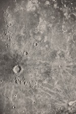 A detailed grayscale image of a lunar surface featuring numerous craters and rugged terrain. The surface is pocked with a variety of circular depressions and raised areas, characteristic of an impact-rich environment. Textured patterns radiate from the largest crater, suggesting past significant impacts.