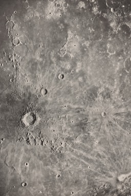 A detailed grayscale image of a lunar surface featuring numerous craters and rugged terrain. The surface is pocked with a variety of circular depressions and raised areas, characteristic of an impact-rich environment. Textured patterns radiate from the largest crater, suggesting past significant impacts.