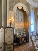 An ornate interior with classical decor, featuring luxurious drapes and detailed wall art depicting cherubs. The room has a sophisticated design with an antique console table adorned with gold candelabras, and a decorative folding screen with floral patterns. Elegant chairs with upholstered seats and intricate carvings are placed alongside the table. Soft lighting from multiple sconces adds a warm glow to the space.