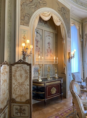 An ornate interior with classical decor, featuring luxurious drapes and detailed wall art depicting cherubs. The room has a sophisticated design with an antique console table adorned with gold candelabras, and a decorative folding screen with floral patterns. Elegant chairs with upholstered seats and intricate carvings are placed alongside the table. Soft lighting from multiple sconces adds a warm glow to the space.