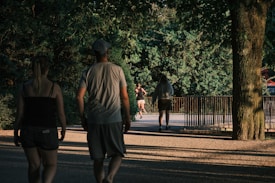 A serene park scene with people walking and jogging along a tree-lined path. The light filtering through the leaves casts soft shadows on the ground. The background features lush green foliage and a metal railing.