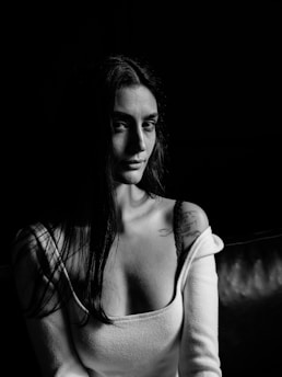 A monochrome portrait of a woman with long hair, wearing an off-the-shoulder top revealing a tattoo on her shoulder. The lighting creates strong contrasts, highlighting her facial features against a dark background.