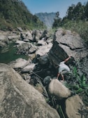 A person navigates across large rocks next to a flowing river, surrounded by dense greenery and towering cliffs in the background. The scene captures an adventurous and natural setting.
