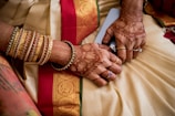 A pair of hands adorned with intricate henna designs and multiple gold and red bangles. The hands rest on a richly decorated fabric featuring a red and gold patterned border.