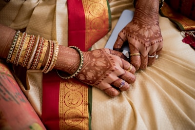 A pair of hands adorned with intricate henna designs and multiple gold and red bangles. The hands rest on a richly decorated fabric featuring a red and gold patterned border.
