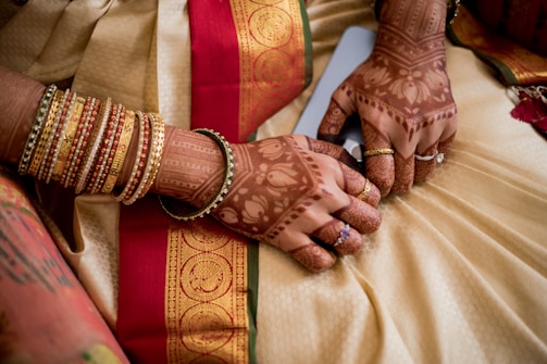 A pair of hands adorned with intricate henna designs and multiple gold and red bangles. The hands rest on a richly decorated fabric featuring a red and gold patterned border.