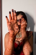 A woman wearing a richly decorated red saree and elaborate jewelry holds her hands in front of her face, showcasing intricate henna designs on her palms and fingers. The henna covers her entire hands and extends to her forearms. Her left eye is visible through her fingers while her right eye is partially covered by her left hand.