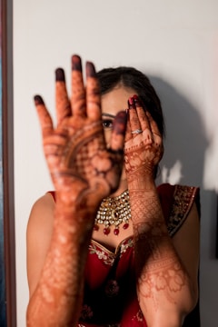 A woman wearing a richly decorated red saree and elaborate jewelry holds her hands in front of her face, showcasing intricate henna designs on her palms and fingers. The henna covers her entire hands and extends to her forearms. Her left eye is visible through her fingers while her right eye is partially covered by her left hand.