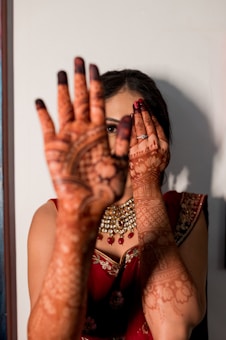 A woman wearing a richly decorated red saree and elaborate jewelry holds her hands in front of her face, showcasing intricate henna designs on her palms and fingers. The henna covers her entire hands and extends to her forearms. Her left eye is visible through her fingers while her right eye is partially covered by her left hand.