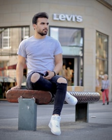 A person in a gray t-shirt and ripped black jeans is sitting on a bench in front of a Levi's store. The individual is wearing white sneakers and holding a phone. The background features storefront windows and a blurred pedestrian.