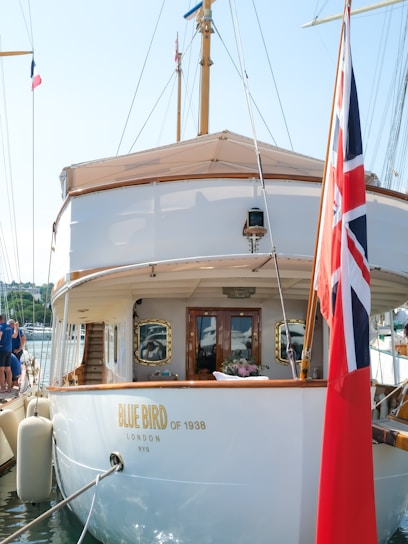 A view of the stern of a classic yacht named 'Blue Bird of 1938' docked at a marina. The yacht is elegantly designed with wooden accents and polished surfaces. The British flag is prominently displayed at the stern, and there are fresh flowers on a table near the door leading into the cabin. Several people in casual attire are seen on the left, likely boarding or attending to another boat.