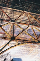 An intricate steel framework with triangular patterns creating a geometric design. The structure features various shades of yellow and orange on the beams, contrasted against a background of a glass or transparent roof.