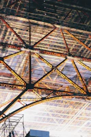 An intricate steel framework with triangular patterns creating a geometric design. The structure features various shades of yellow and orange on the beams, contrasted against a background of a glass or transparent roof.
