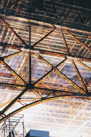 An intricate steel framework with triangular patterns creating a geometric design. The structure features various shades of yellow and orange on the beams, contrasted against a background of a glass or transparent roof.