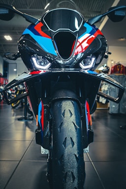 A close-up view of a high-performance motorcycle featuring an aggressive front design with sharp headlights and a colorful body with black, blue, red, and orange details. The image captures the bike's front tire, aerodynamic components, and a part of the showroom in the background.