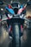 A close-up view of a high-performance motorcycle featuring an aggressive front design with sharp headlights and a colorful body with black, blue, red, and orange details. The image captures the bike's front tire, aerodynamic components, and a part of the showroom in the background.