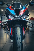 A close-up view of a high-performance motorcycle featuring an aggressive front design with sharp headlights and a colorful body with black, blue, red, and orange details. The image captures the bike's front tire, aerodynamic components, and a part of the showroom in the background.