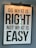 A framed motivational poster with the phrase 'Do what is right, not what is easy' written in large, bold black lettering on a white background. A light fixture is positioned above the frame, casting a warm glow. Reflections of a window or similar structures are visible on the surface of the glass.