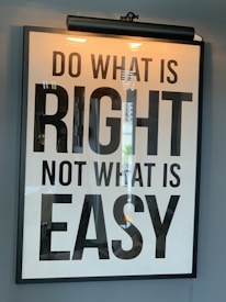 A framed motivational poster with the phrase 'Do what is right, not what is easy' written in large, bold black lettering on a white background. A light fixture is positioned above the frame, casting a warm glow. Reflections of a window or similar structures are visible on the surface of the glass.