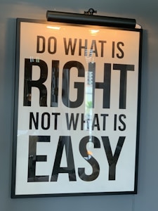 A framed motivational poster with the phrase 'Do what is right, not what is easy' written in large, bold black lettering on a white background. A light fixture is positioned above the frame, casting a warm glow. Reflections of a window or similar structures are visible on the surface of the glass.