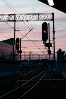 A railway track is silhouetted against a pastel-colored sunset sky. Two red-lit signals stand prominently over the tracks, with overhead wires and support structures creating an intricate network above the railway. In the background, the outline of buildings and more railway infrastructure can be seen.