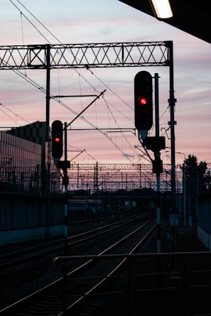 A railway track is silhouetted against a pastel-colored sunset sky. Two red-lit signals stand prominently over the tracks, with overhead wires and support structures creating an intricate network above the railway. In the background, the outline of buildings and more railway infrastructure can be seen.