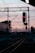 A railway track is silhouetted against a pastel-colored sunset sky. Two red-lit signals stand prominently over the tracks, with overhead wires and support structures creating an intricate network above the railway. In the background, the outline of buildings and more railway infrastructure can be seen.