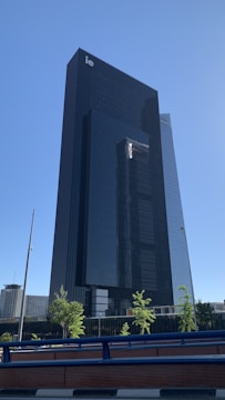 A modern skyscraper with a sleek, dark glass facade stands tall against a clear blue sky. There are a few small trees and a railing in the foreground. The design is minimalist and contemporary, showcasing vertical lines and reflective surfaces.