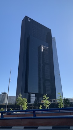 A modern skyscraper with a sleek, dark glass facade stands tall against a clear blue sky. There are a few small trees and a railing in the foreground. The design is minimalist and contemporary, showcasing vertical lines and reflective surfaces.