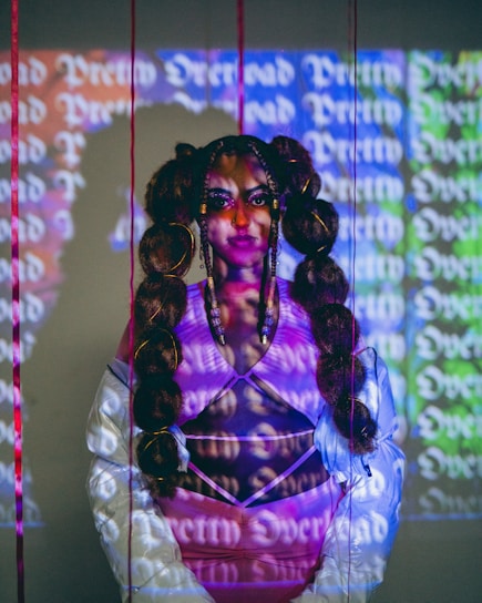 A person with intricate braided hair stands in front of a background featuring colorful, repetitive text. The person is wearing a white jacket with a pink top underneath, and their face is illuminated by the text projected onto them.