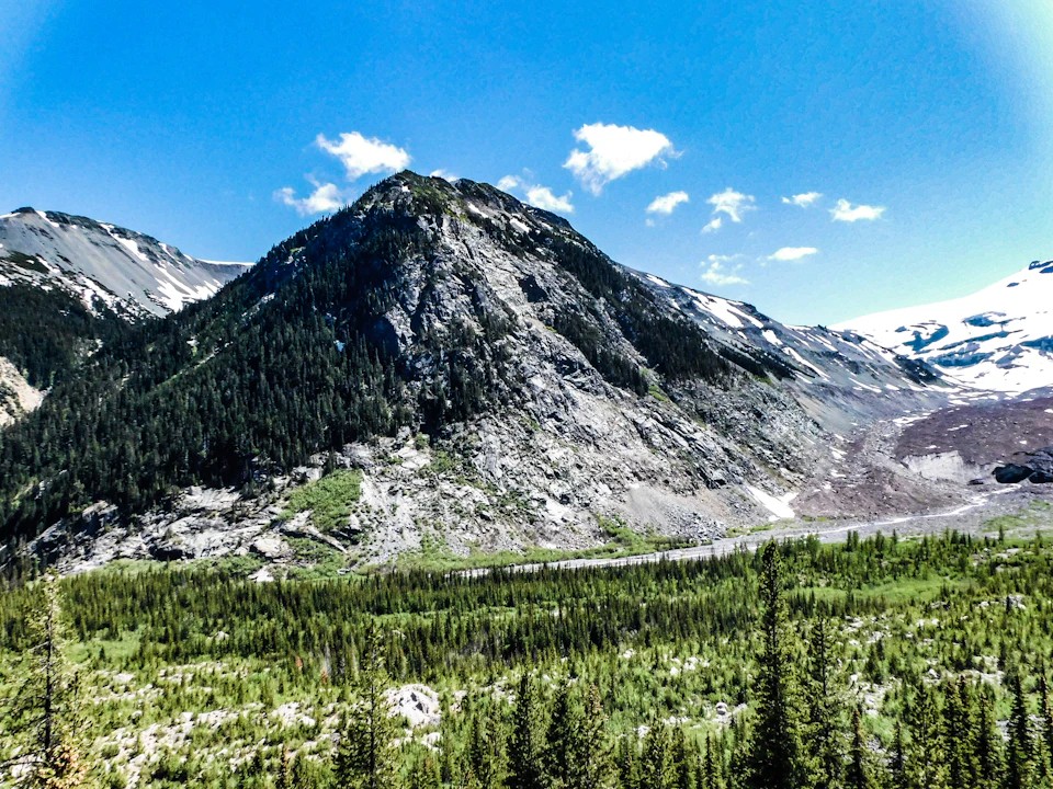 The image depicts a breathtaking mountain landscape with a large, jagged gray rock formation on the left side and a valley filled with lush green trees on the right side. The sky above is a clear blue, dotted with fluffy white clouds. The perspective of the photo suggests it was taken from a high vantage point, providing an expansive view of the surrounding mountains and valleys.