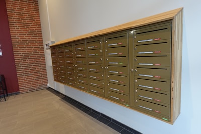 A wall-mounted array of metallic mailboxes with wooden framing, featuring multiple horizontal slots for mail. Each mailbox slot has a label with red or green indicators. The background includes a partial brick wall alongside a light-colored painted wall and tiled floor.