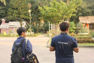 Two individuals are walking in an outdoor area with trees and small structures in the background. They are wearing navy shirts with 'TRUE HAPPINESS' written on the back, each carrying a backpack. The setting has a casual and relaxed atmosphere.
