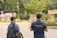 Two individuals are walking in an outdoor area with trees and small structures in the background. They are wearing navy shirts with 'TRUE HAPPINESS' written on the back, each carrying a backpack. The setting has a casual and relaxed atmosphere.