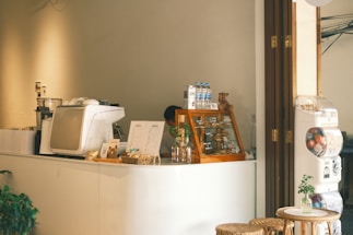 A cozy cafe counter area featuring a white espresso machine, a display case with baked goods, and a variety of items including bottled water and a small teddy bear. Menu cards are prominently displayed on the counter. To the side, there is a vending machine filled with colorful capsules. The setting includes wooden accents, potted plants, and a minimalist decor style.