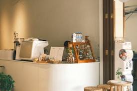 A cozy cafe counter area featuring a white espresso machine, a display case with baked goods, and a variety of items including bottled water and a small teddy bear. Menu cards are prominently displayed on the counter. To the side, there is a vending machine filled with colorful capsules. The setting includes wooden accents, potted plants, and a minimalist decor style.