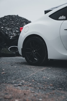A close-up of the rear section of a sleek white car parked on a rough, gravelly surface. The car features black wheels and a visible taillight. In the background, a pile of tires or circular objects can be seen, contributing to an industrial atmosphere.