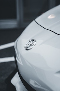 A close-up view of the front of a white automobile featuring the Toyota logo prominently displayed on the hood. The image captures the sleek and smooth lines of the vehicle against a blurred background.