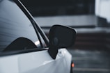 Close-up of a car's side mirror and window against a blurred, dark background. The vehicle appears sleek with a glossy white exterior.