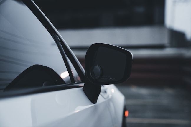 Close-up of a car's side mirror and window against a blurred, dark background. The vehicle appears sleek with a glossy white exterior.